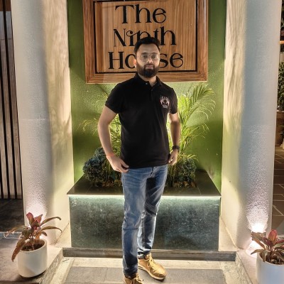 Headshot of Sumit Kumar, AVP Product at Statiq, standing in front of a wooden sign reading The Ninth House, wearing a black polo shirt and jeans