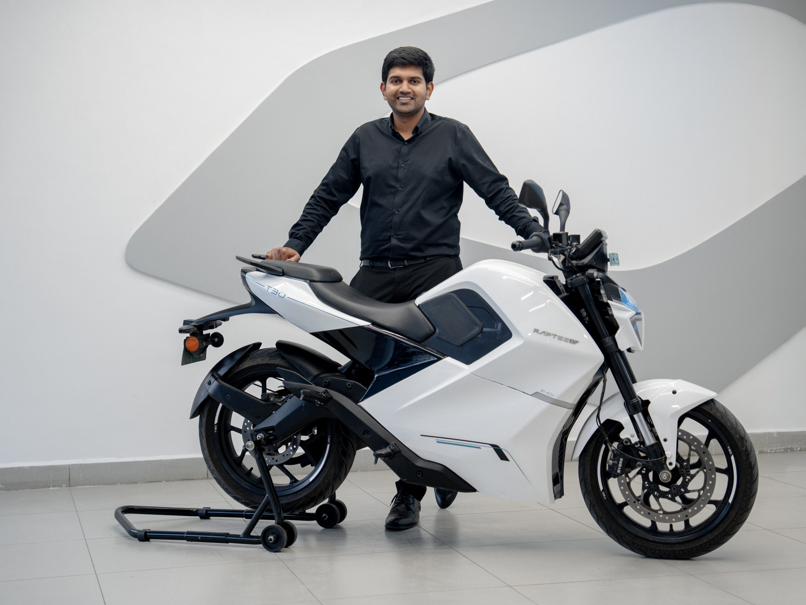 Headshot of Dinesh Arjun, Co-founder and CEO of Raptee.HV, smiling in a black shirt beside a white electric motorcycle