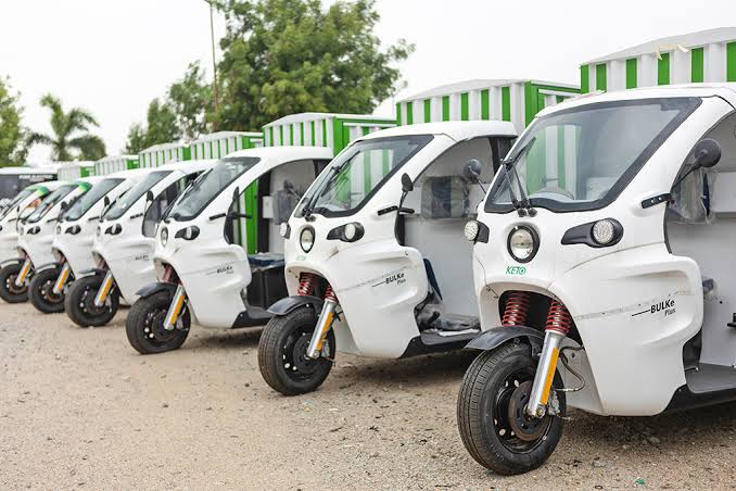 White ETO three-wheeler with a cargo body and green stripes beside an ETO Thunderbox charging unit on a city-skyline plaza