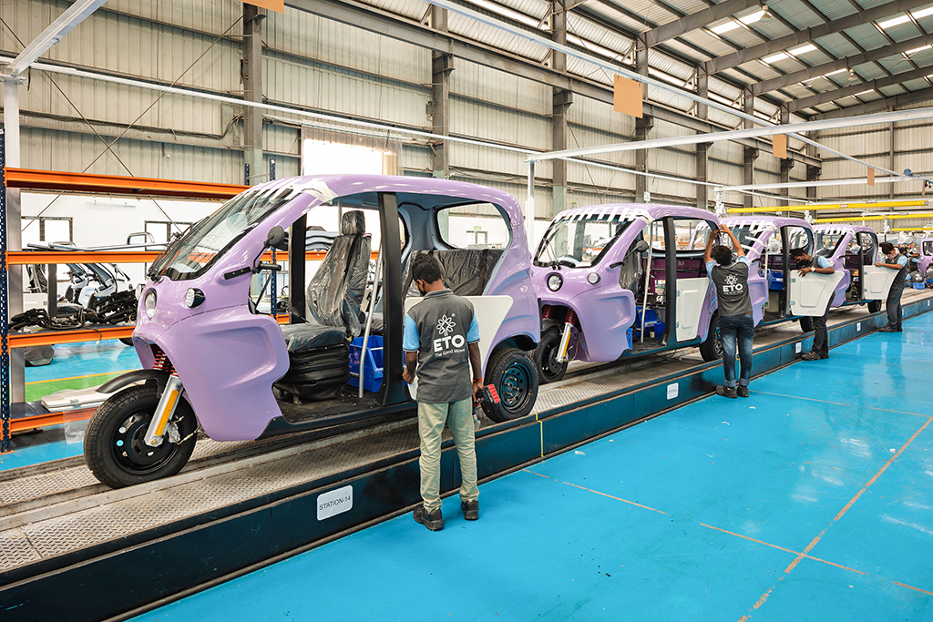 Five men in white safety helmets walking along an assembly floor lined with parts racks, led by a man in a blue shirt and followed by colleagues in ETO uniform