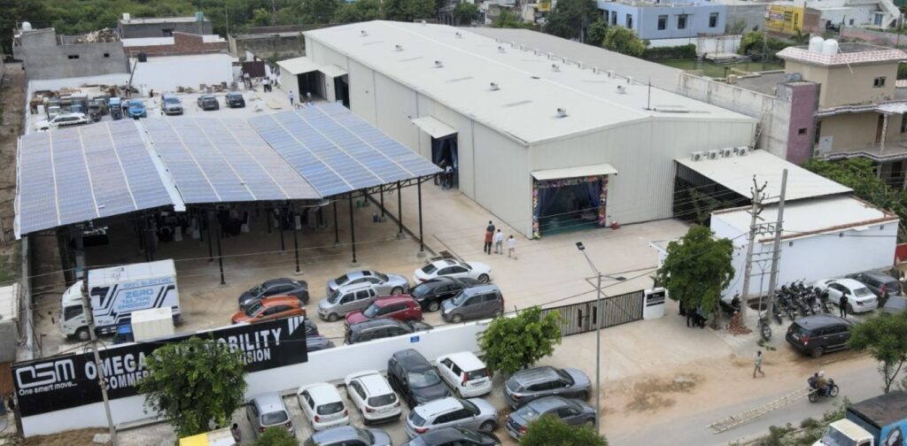 Omega Seiki Mobility manufacturing facility with a large white industrial shed, a solar-roofed parking canopy and a black OSM Omega Seiki Mobility signboard in front of the plant