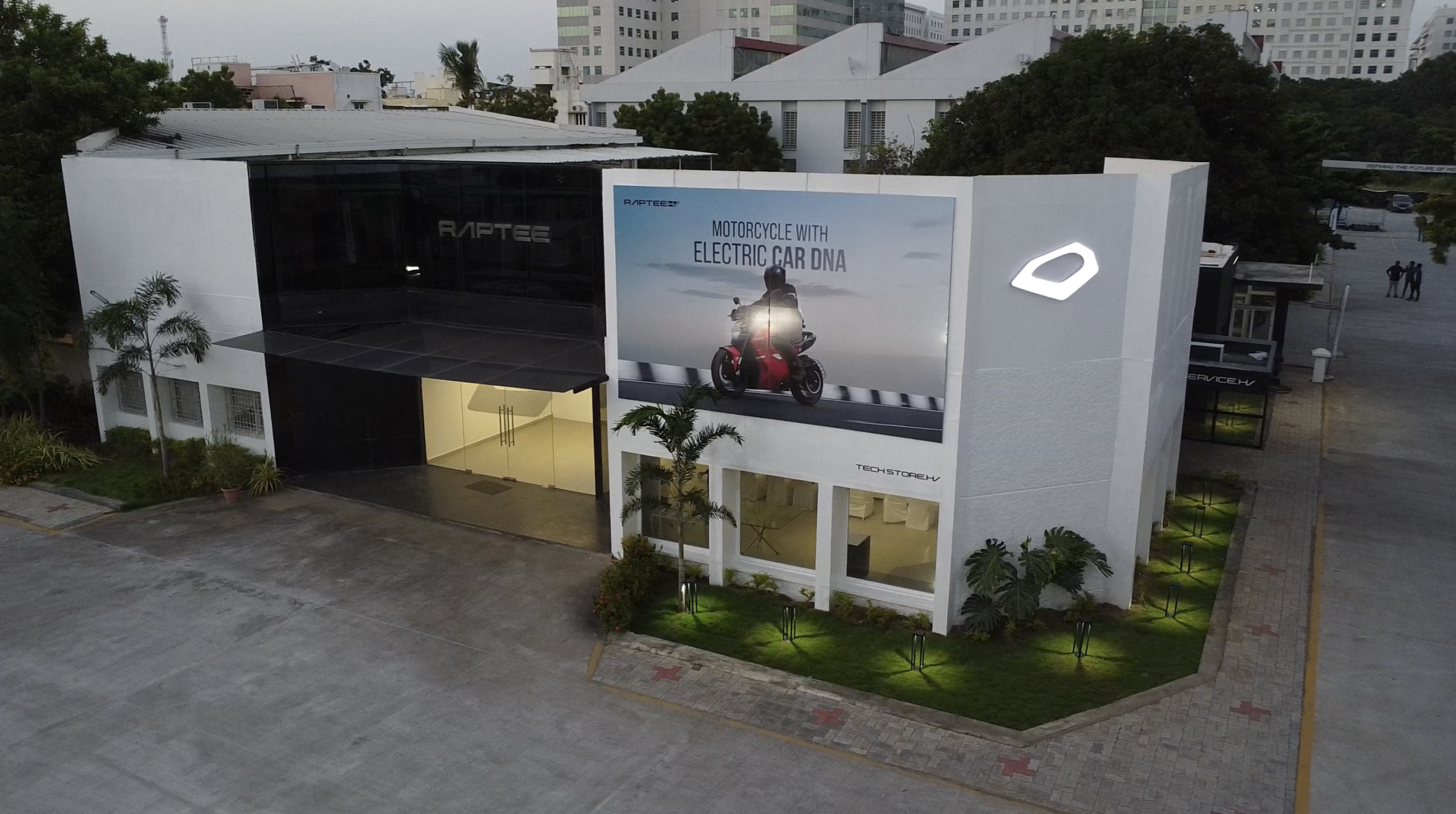 Raptee.HV tech store building with a white facade, illuminated logo sign and a billboard reading Motorcycle with Electric Car DNA, set among trees in an urban campus